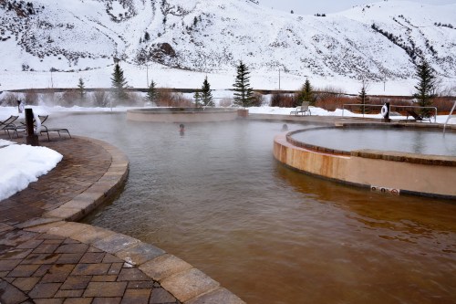 A serene hot spring surrounded by snow-covered mountains, with mist rising from the warm water.