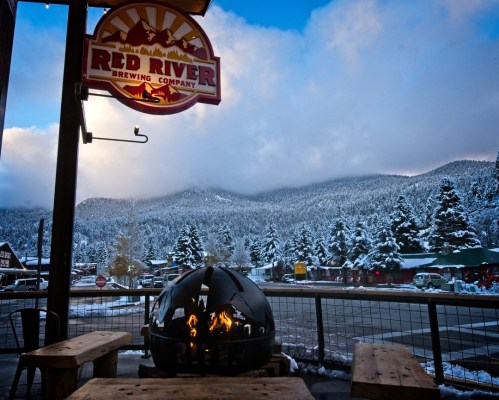 A cozy outdoor scene featuring a fire pit, wooden tables, and a sign for Red River Brewing Company against a snowy mountain backdrop.