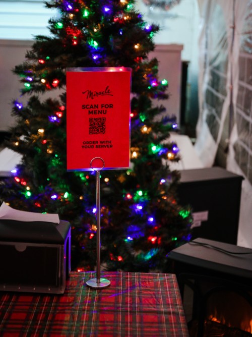 A festive table with a sign to scan for the menu, next to a decorated Christmas tree with colorful lights.