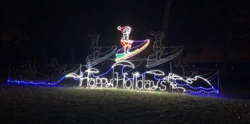 Colorful holiday lights display featuring a surfing figure and the text "Happy Holidays" on a wave backdrop.