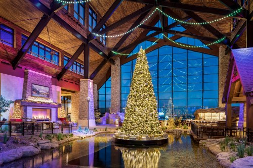 A beautifully decorated Christmas tree stands in a cozy lodge with a large window showcasing a nighttime city view.