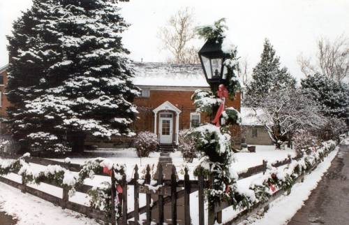 A snowy scene of a brick house with a festive fence, decorated with garlands and a lamp post adorned with a bow.