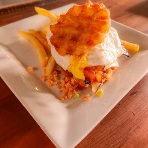 A plate of fries topped with a fried egg and a crispy waffle, served on a white square dish.