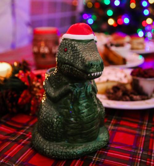 A festive dinosaur figurine wearing a Santa hat, surrounded by holiday treats and colorful lights.