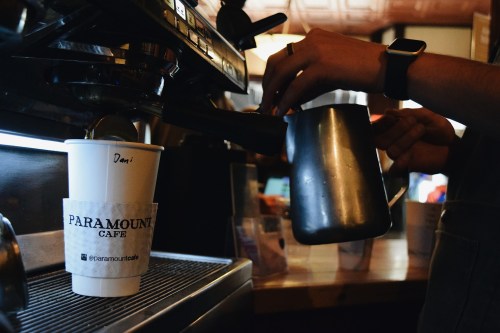 A barista pours steamed milk into a coffee cup at Paramount Cafe, with a focus on the cup's branding.
