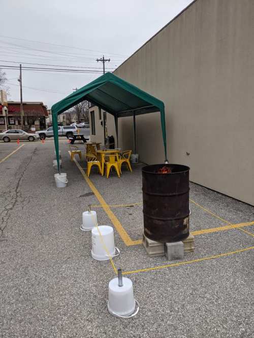 A green tent with yellow chairs and a barrel fire in a parking lot, surrounded by white buckets.