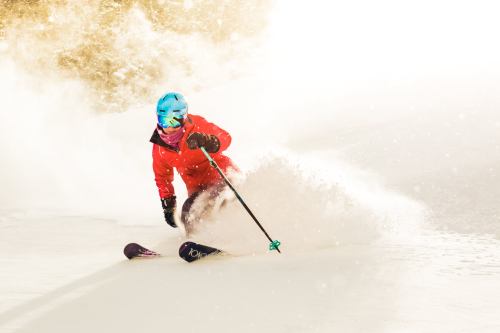 A skier in a red jacket carves through fresh snow, creating a spray of powder against a bright background.