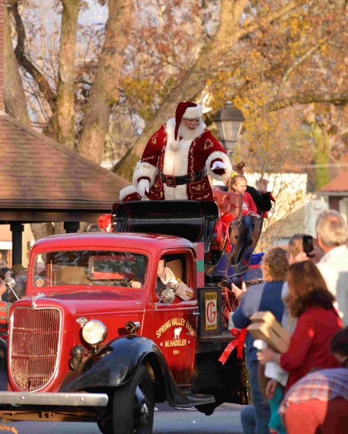 A vintage red truck with Santa and children, parading through a festive crowd in a park setting.