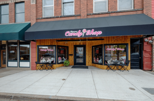 Exterior of a candy shop with a black awning, large windows, and outdoor seating on a city sidewalk.