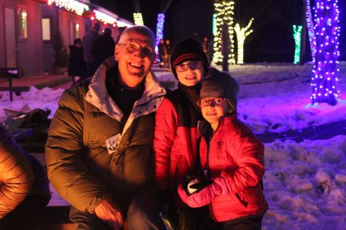 A smiling man sits with two children in winter attire, surrounded by colorful holiday lights and snow.