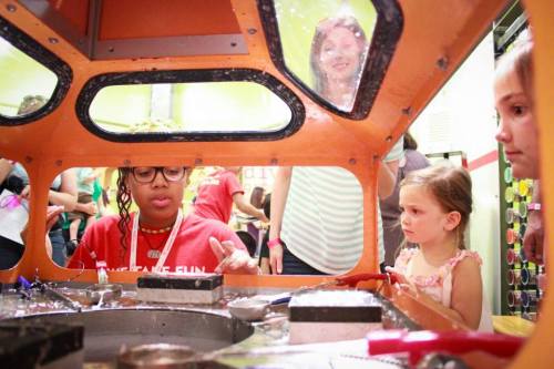 A child watches as an educator demonstrates an activity inside a colorful exhibit, with other visitors in the background.