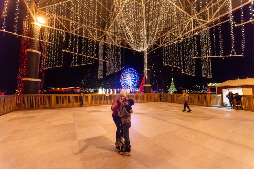 Two people embrace on a festive ice rink, surrounded by colorful lights and a Ferris wheel in the background.