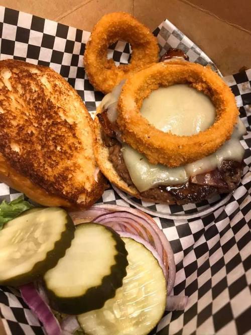 A delicious burger with melted cheese, bacon, and onion rings, served with pickles and red onion on a checkered paper.