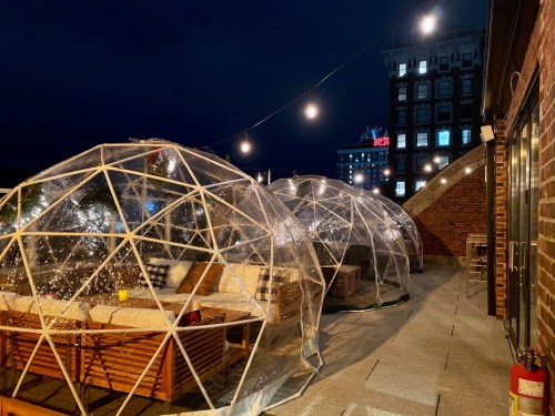 A rooftop terrace with transparent igloo-like structures, cozy seating, and string lights against a night sky.
