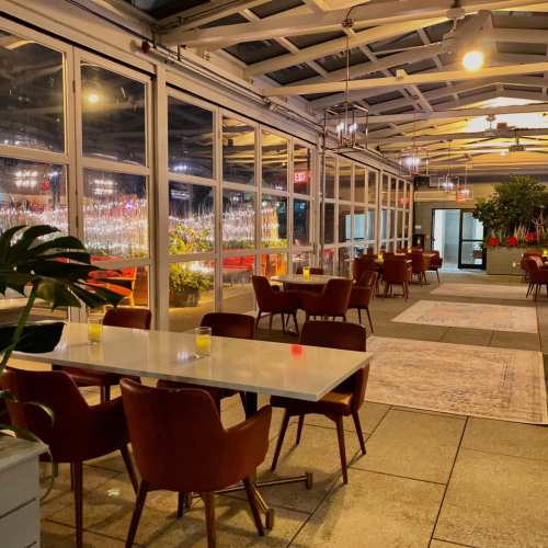 Cozy restaurant interior with glass walls, warm lighting, and modern furniture, featuring empty tables and plants.