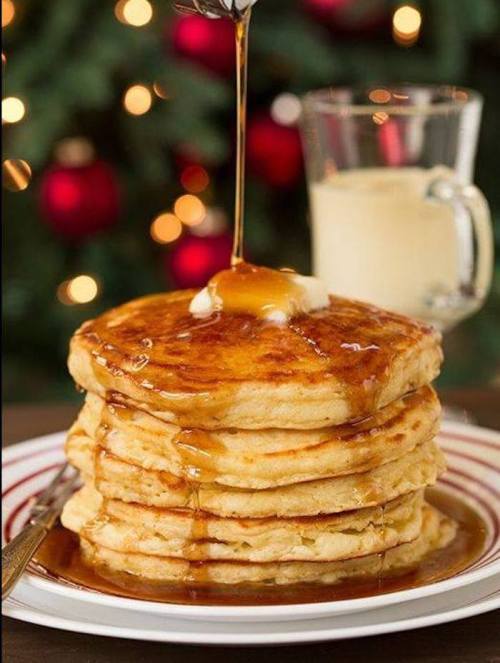A stack of fluffy pancakes drizzled with syrup, topped with butter, beside a glass of milk and a festive background.