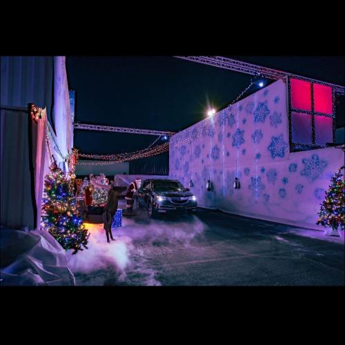 A festive outdoor scene with decorated trees, a car, and snowflake projections, illuminated by colorful lights at night.