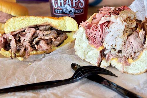 A close-up of two hearty sandwiches filled with various meats, served with a black fork on a white paper.