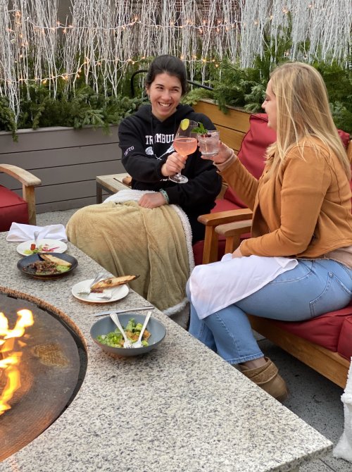 Two women toast with drinks while sitting by a fire pit, enjoying food on a cozy outdoor patio.