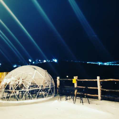 A clear igloo-shaped structure sits on snow, with a view of distant lights and a wooden railing at night.