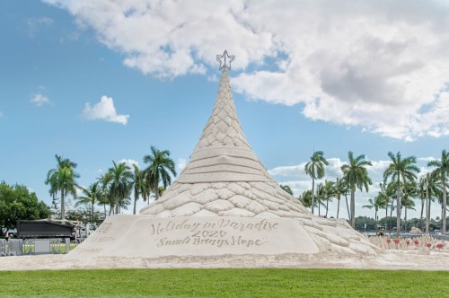 A large sand sculpture of a Christmas tree with a star on top, surrounded by palm trees and a blue sky.