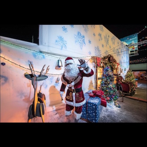 A festive scene featuring Santa Claus waving, surrounded by holiday decorations, a Christmas tree, and gifts.