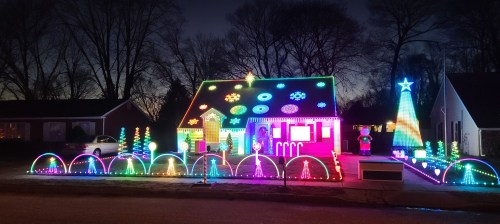 A brightly lit house decorated with colorful Christmas lights, featuring trees, snowflakes, and a star, at dusk.