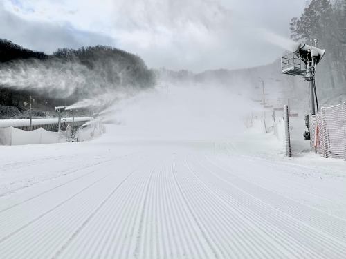 Snowmaking machines create a snowy landscape on a ski slope, with freshly groomed trails and misty snow in the air.