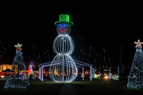A large illuminated snowman with a green hat, surrounded by glowing Christmas trees and festive lights at night.