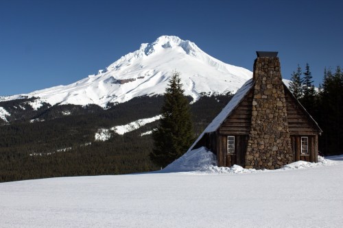 A rustic cabin with a stone chimney in a snowy landscape, with a majestic snow-capped mountain in the background.