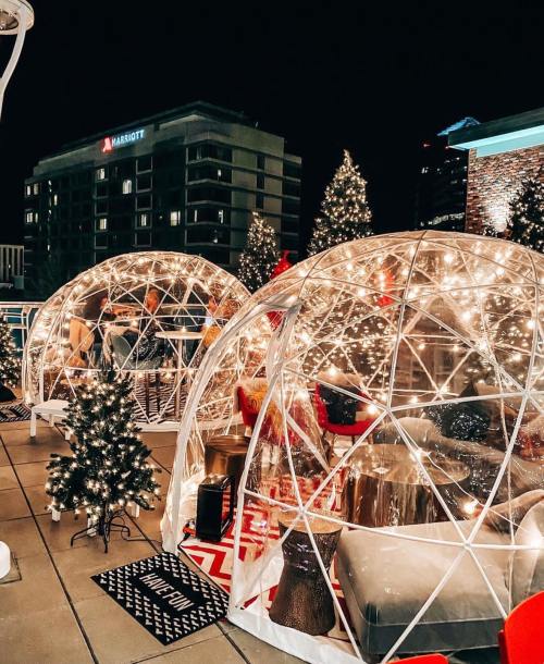 Two illuminated igloos on a rooftop, surrounded by Christmas trees and festive decorations at night.