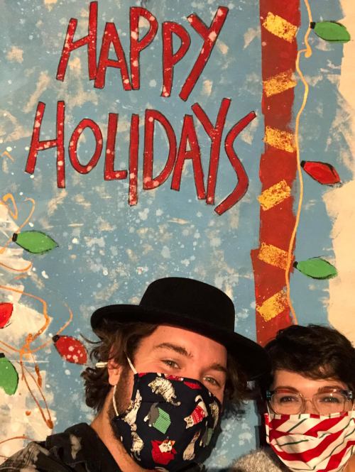 Two people wearing festive masks pose in front of a colorful "Happy Holidays" backdrop with decorations.