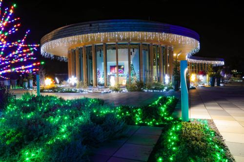 A circular building adorned with colorful holiday lights, surrounded by green illuminated landscaping at night.
