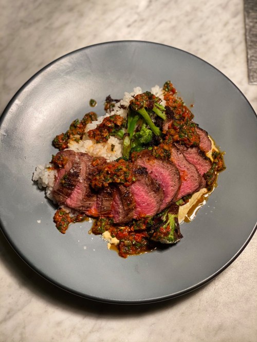 Sliced steak on a bed of rice, topped with vibrant green sauce and garnished with broccoli on a dark plate.