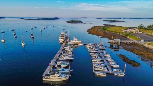 Aerial view of a marina with boats docked, surrounded by calm water and distant islands under a clear blue sky.