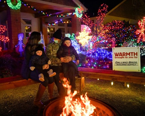 A family sits by a fire pit, enjoying colorful holiday lights in the background, with a sign reading "WARMTH."