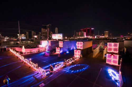 A festive outdoor scene at night, featuring colorful lights, decorations, and large gift boxes in an urban setting.