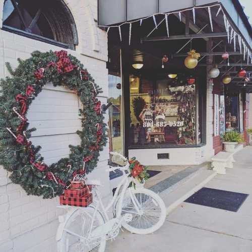 A festive storefront decorated with a wreath, holiday ornaments, and a white bicycle adorned with flowers and gifts.