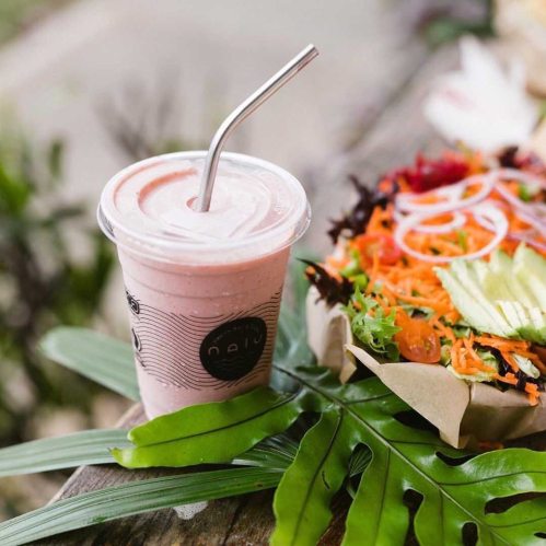 A smoothie in a cup with a straw next to a colorful salad, surrounded by green leaves.