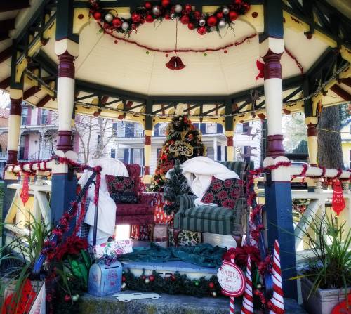 A decorated gazebo with a Christmas tree, two cozy chairs, and festive ornaments, surrounded by holiday decorations.