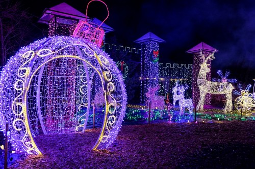 Colorful holiday lights illuminate a festive scene with a large ornament, reindeer, and a castle backdrop at night.