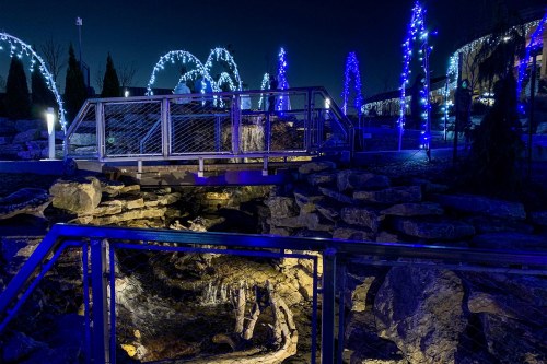 A nighttime scene featuring a bridge over rocks, illuminated by blue lights and surrounded by festive decorations.