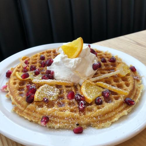A golden waffle topped with whipped cream, orange slices, and pomegranate seeds, served on a white plate.
