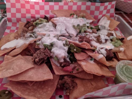 A tray of nachos topped with meat, guacamole, jalapeños, and drizzled with sour cream, served on a checkered paper.