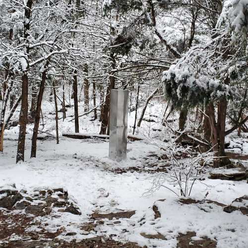 A tall, reflective monolith stands in a snowy forest surrounded by trees and a snowy path.