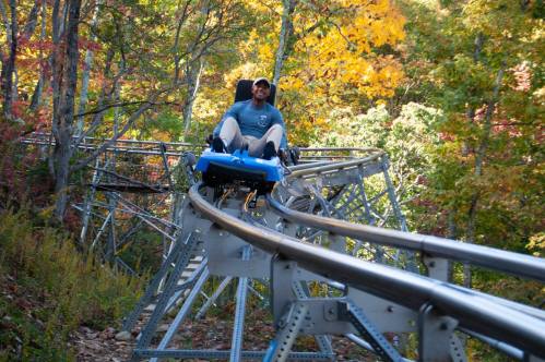 A person rides a blue alpine coaster on a metal track surrounded by colorful autumn trees.