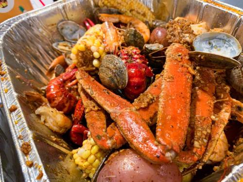 A metal tray filled with a variety of seafood, including crab, shrimp, clams, and corn, mixed with spices.