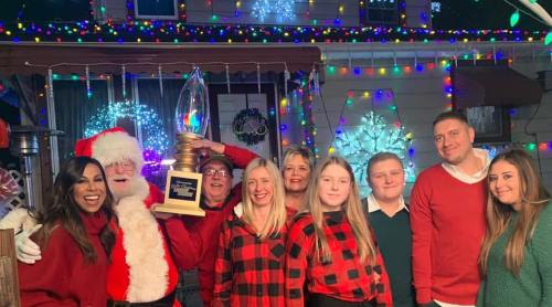 A festive group poses with Santa in front of a brightly decorated house for the holidays, holding a trophy.