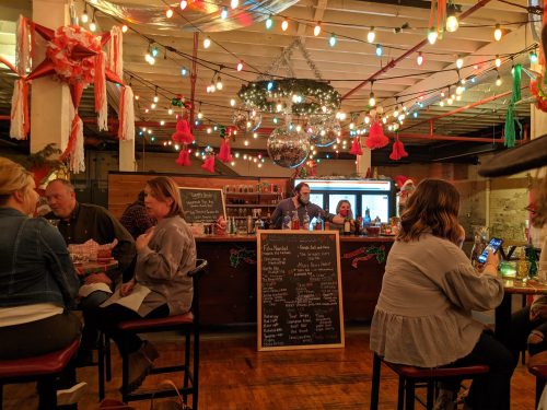 A festive bar scene with colorful lights, patrons enjoying drinks, and a bartender behind a counter decorated for the holidays.