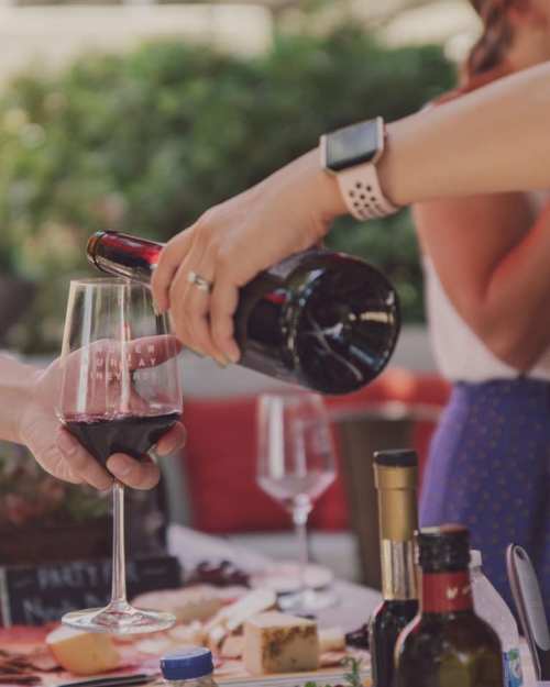 A hand pours red wine into a glass at a table with various food items and drinks in a relaxed outdoor setting.
